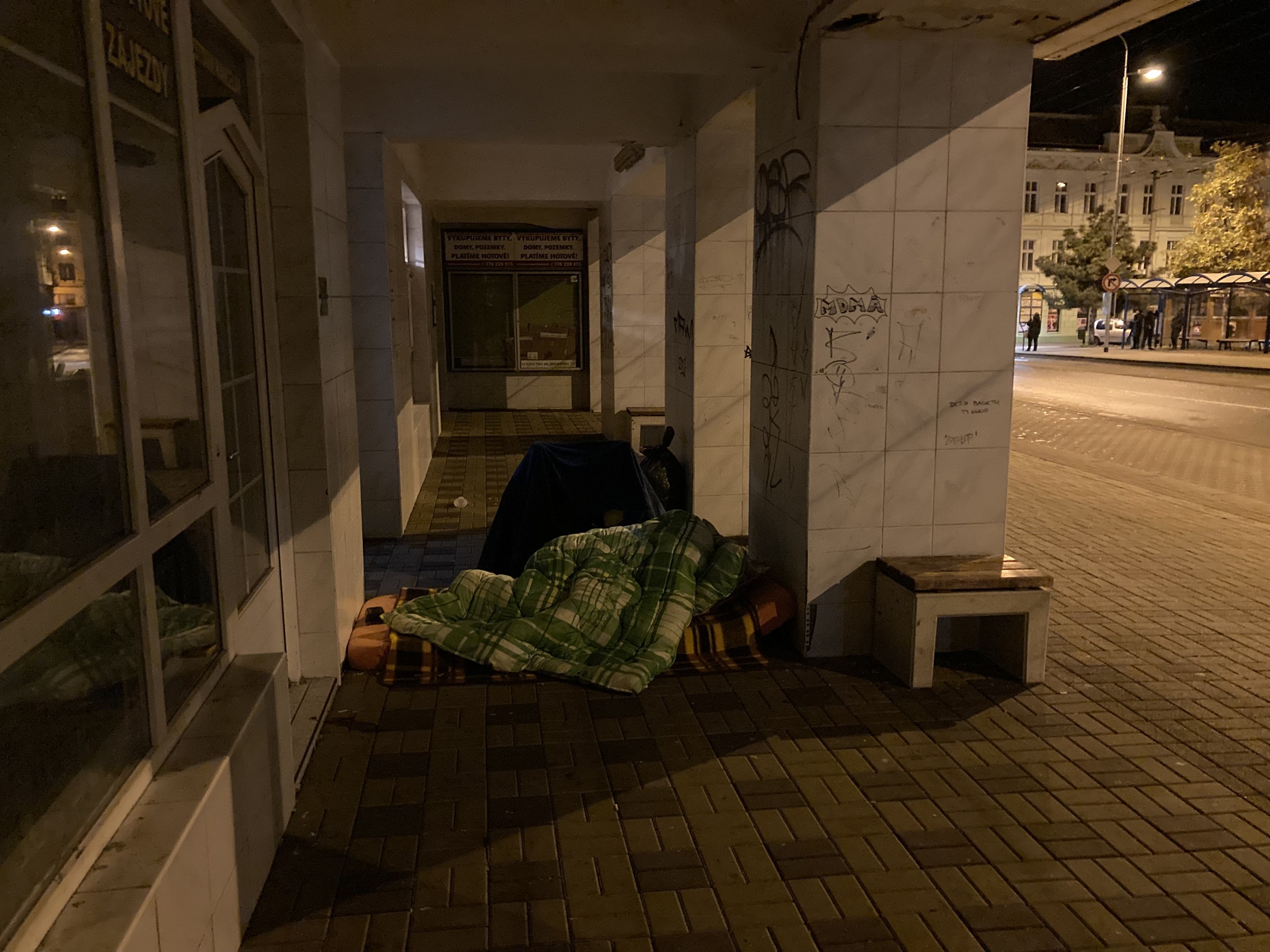 Eine obdachlose Person, die nachts auf einem Bürgersteig schläft, mit einer Decke bedeckt, mit einer Bank in der Nähe, umgeben von Menschen, Fahrzeugen, einem Straßenpfahl, Bäumen, einem Schild, Gebäuden mit Fenstern und einem bewölkten Himmel.