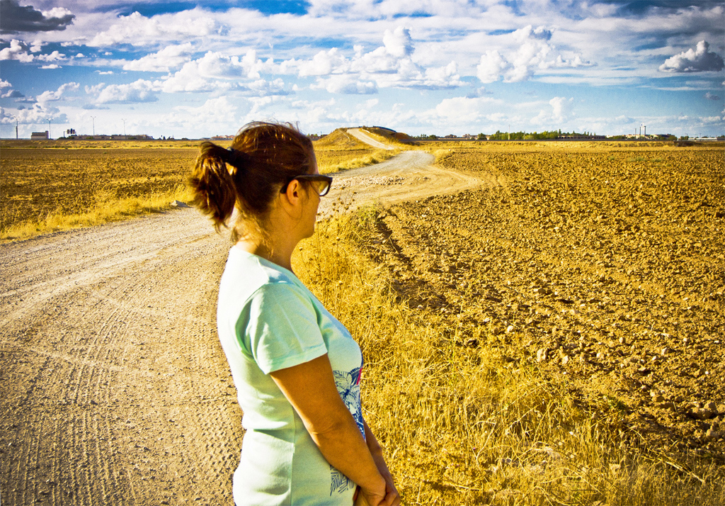 Eine Frau steht auf einem schlammigen Weg, flankiert von Landwirtschaftsland und Bäumen.