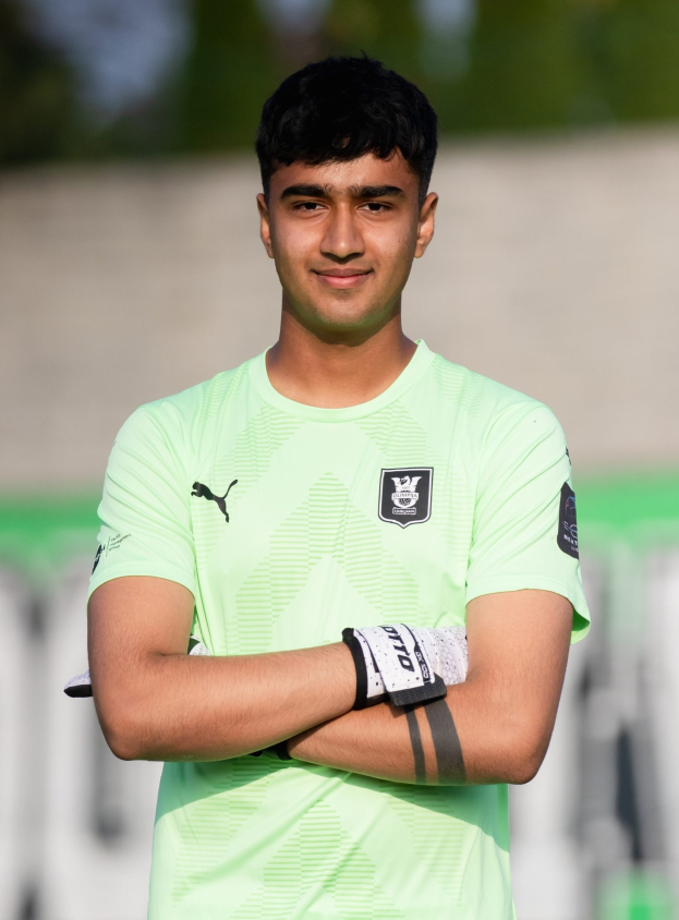 Ein junger Mann in einem grünen Hemd steht auf einem Fußballfeld und hat die Arme verschränkt, während er lächelt, mit einem unscharfen Hintergrund.