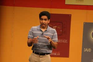 Ein Mann steht auf einer Bühne und spricht mit einer Wand voller Plakate dahinter.