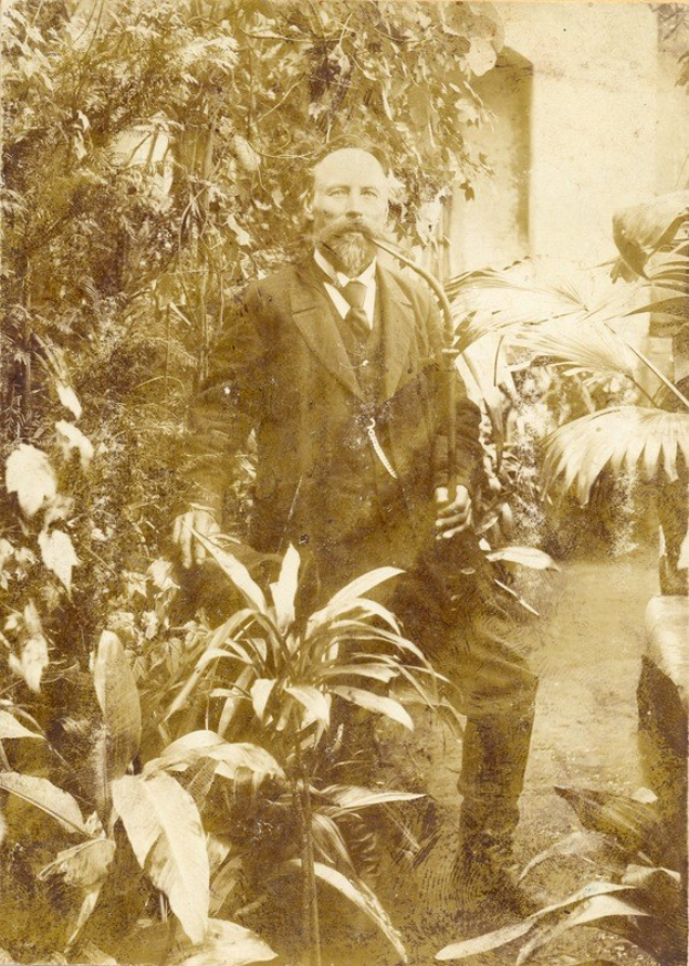 Altes Foto eines Mannes in Anzug und Krawatte, der in einem Garten mit Pflanzen und Bäumen steht, eine Wand ist im Hintergrund sichtbar.