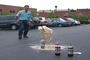 Ein Mann steht auf einer Straße, lacht, mit drei Flaschen vor ihm, während Autos, eine Pflanze, ein Lichtmast und der Himmel im Hintergrund zu sehen sind.