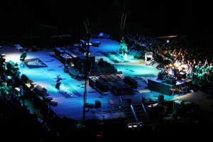 Menschen, die auf einer Bühne mit Boxen, Lautsprechern, Lampen und Mikrofonen stehen, mit einer großen Menge auf beiden Seiten und einem dunklen Hintergrund.