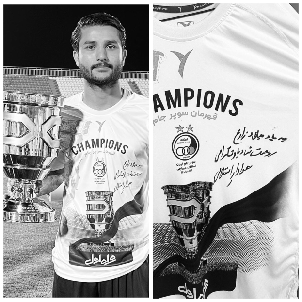 Ein Mann in einem weißen 'Champions'-T-Shirt hält einen Pokal, während er in einem Stadion steht.