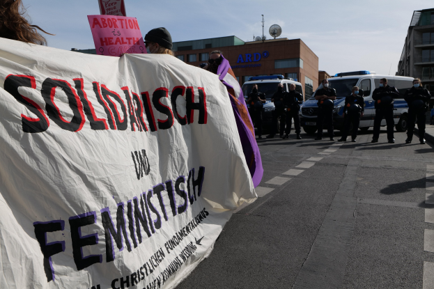 Eine Gruppe von Menschen marschiert auf der Straße und hält ein Banner mit der Aufschrift "Solidarität und Feminismus", mit parkenden Fahrzeugen, Gebäuden, einer Schüsselantenne und einem klaren blauen Himmel im Hintergrund.
