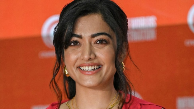 Eine Frau in einem roten Kleid mit einem eleganten Dutt und dezentem Make-up lächelt in die Kamera, trägt schwarze High Heels und hat ein hellgelbes Banner mit der Aufschrift "Sonam Kapoors beste Beauty-Looks vom Cannes Film Festival" im Hintergrund.