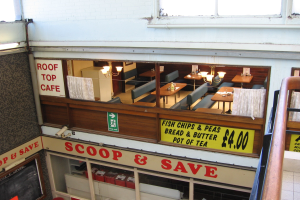 Ein café-ähnliches Restaurant mit Tischen, Stühlen und beleuchteten Tafeln mit Text, das eine Brüstung auf der rechten Seite und eine Wand im Hintergrund mit der Aufschrift 'Scoop & Save' hat.