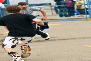 Zwei Personen spielen Hockey auf einer Straße mit einem Zaun dahinter und einer Menge von Menschen im Hintergrund.