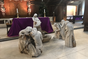 Krippe in einer Kirche mit Statuen auf dem Boden, einem Tisch mit einer Decke und Kerzenständern, Stühlen, einem geschmückten Weihnachtsbaum und einer Wand im Hintergrund.