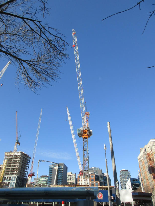 Eine Gruppe von Baukränen auf einer Straße umgeben von Gebäuden, Bäumen, Masten, Brettern und anderen Gegenständen mit dem Himmel im Hintergrund.