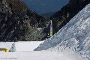 Ein Mann in Skier springt in die Luft mit schneebedeckten Bergen und einer Flagge mit Text im Hintergrund und einem anderen Mann vor der Flagge mit Text in der linken unteren Ecke.