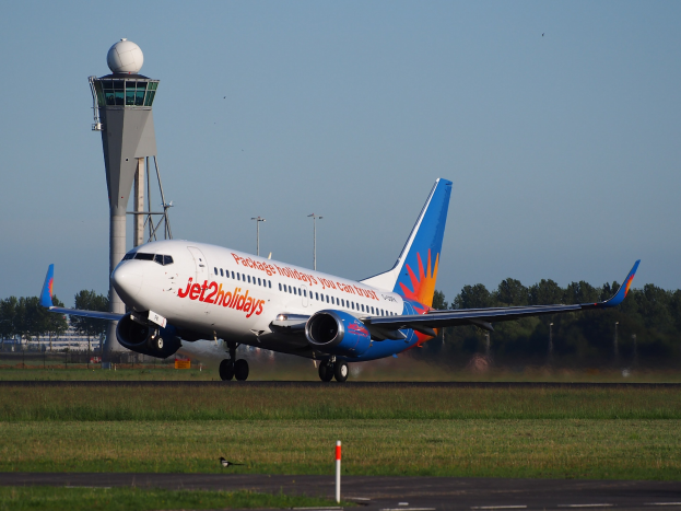 Ein Airbus A320-200 der Jet2holidays von Frankfurt Airport abhebend, umgeben von grünem Gras und einem hohen Turm im Hintergrund, mit Bäumen und Gebäuden in der Ferne unter einem hellblauen Himmel.