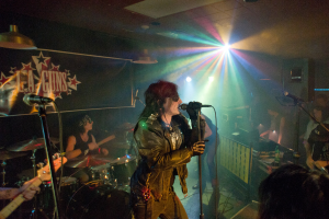 Eine Frau in einer schwarzen Jacke singt in ein Mikrofon auf der Bühne, mit einem Mann in Sonnenbrille und einem Stirnband, der ein Instrument spielt, und einem Plakat mit 'LA Guns' im Hintergrund.