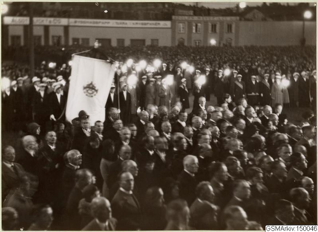 Schwarzes und weißes Bild einer großen Menschenmenge vor einem Gebäude, mit einer Person, die eine Fahne hält, in der Mitte, Stangen und Lichter im Hintergrund und Text unten, der "Gustav Klimts Rede an der Berliner Mauer" lautet.
