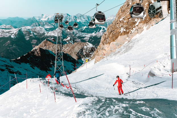 Gruppe von Menschen, die eine schneebedeckte Abfahrt mit einem Seilbahnzug im Hintergrund hinunterfahren, unter einem klaren blauen Himmel mit einer Bergkette.