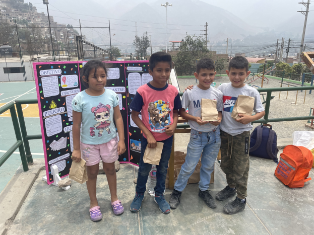 Gruppe von Kindern auf einem Basketballfeld mit Papierbeuteln in den Händen, mit Gegenständen auf der rechten Seite und einer Tafel mit Text auf der linken Seite, vor einem Hintergrund aus Zäunen, Pfosten, Straßenlaternen, Bäumen, Gebäuden, Hügeln und Himmel.