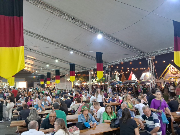 Eine große Gruppe von Menschen sitzt an Tischen unter einem Zelt auf dem Oktoberfest in München, Deutschland, mit Taschen und Gegenständen auf den Tischen, umgeben von festlichen Zelten, Lichtern und Fahnen.