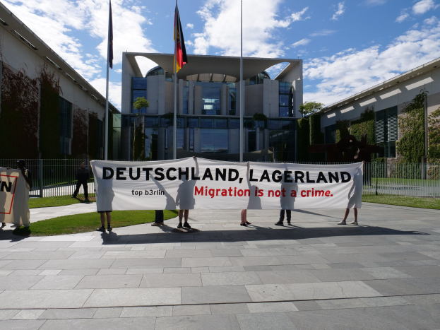 Gruppe von Menschen mit einer Fahne mit der Aufschrift "Deutschland, Lagerland Migration ist kein Verbrechen" vor einem Gebäude mit Fenstern, umgeben von Gras, einem Metallzaun, Pflanzen, Bäumen und Fahnenmasten unter einem bewölkten Himmel.