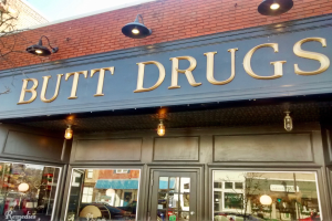 Ein Gebäude mit einer Leuchtschrift, die 'butt drugs' anzeigt, umgeben von Glaswänden und -türen, mit sichtbaren Fahrzeugen, Gebäuden, Bäumen und Himmel im Inneren.