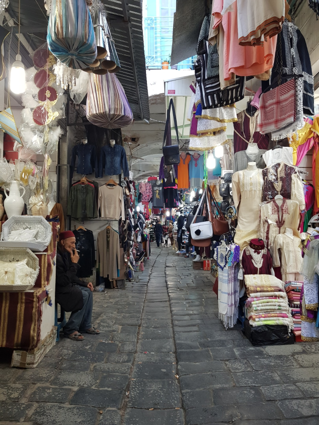 Ein belebter Straßenmarkt in der Altstadt von Jerusalem mit Ständen, die Kleidung, Taschen, Lampen und andere Artikel verkaufen, umgeben von Menschen, die durch die Stände schlendern und ein Gebäude im Hintergrund.