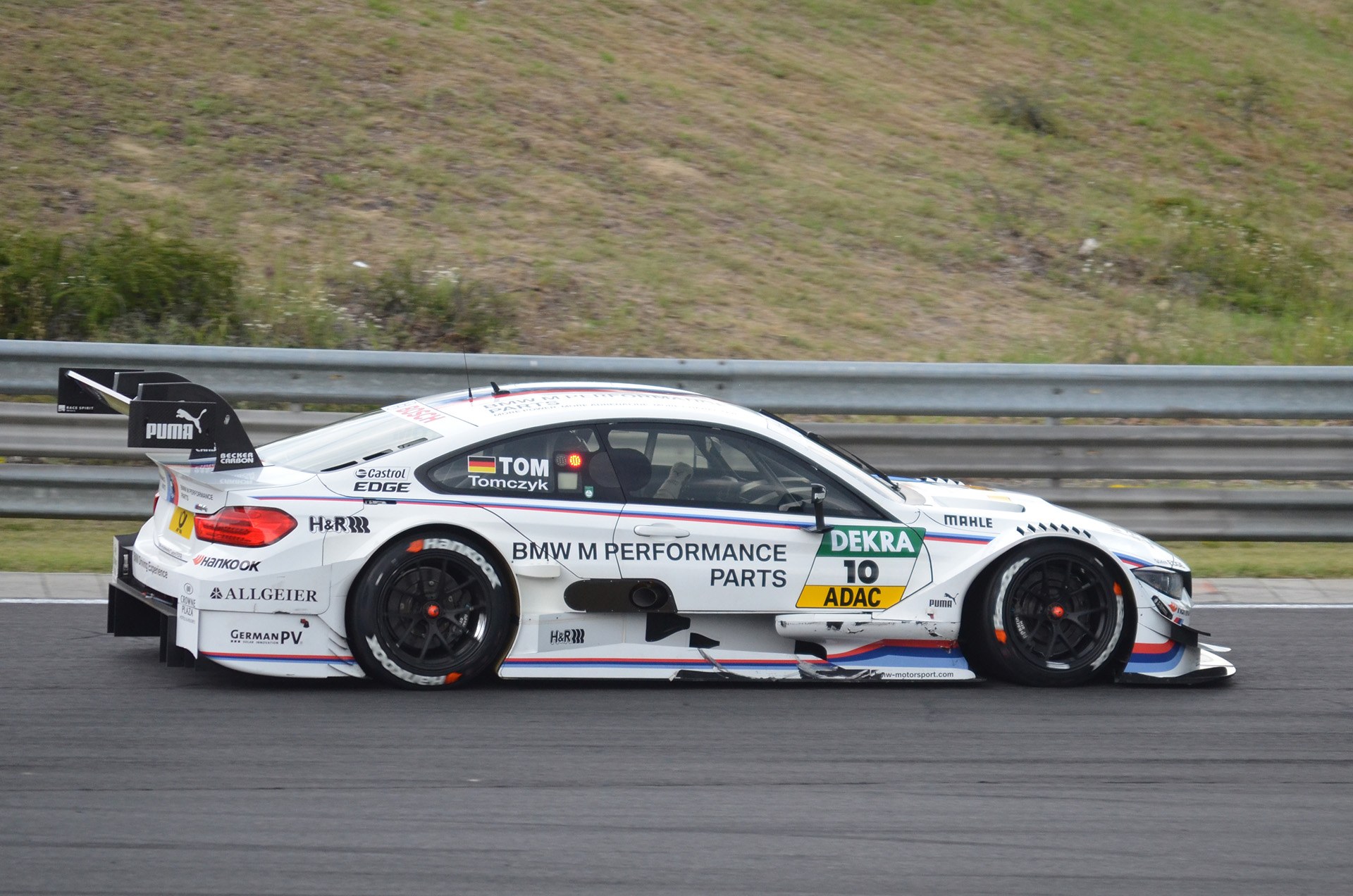 BMW M4 DTM-Rennwagen auf einer Rennstrecke mit Werbetext, umgeben von grünem Gras und einer Metallabsperrung im Hintergrund.