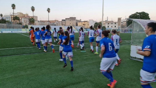Eine Gruppe von Frauen, die auf einem Feld Fußball spielen, mit Gebäuden, Bäumen und einem Torpfosten im Hintergrund.