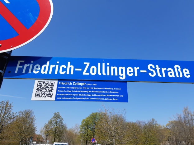 Straßenschild mit einem QR-Code davor vor einem Gebäude, mit Fahrzeugen, Bäumen und einem klaren blauen Himmel im Hintergrund.