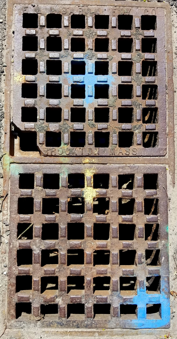 Metallschachtdeckel mit Gittermuster an der Seite eines Gebäudes.