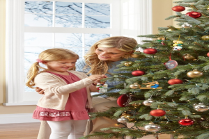 Ein geschmückter Weihnachtsbaum steht in einem Haus mit einer Frau und einem Kind im Vordergrund, einem Fenster und einer Wand im Hintergrund und Bäumen vor dem Fenster.