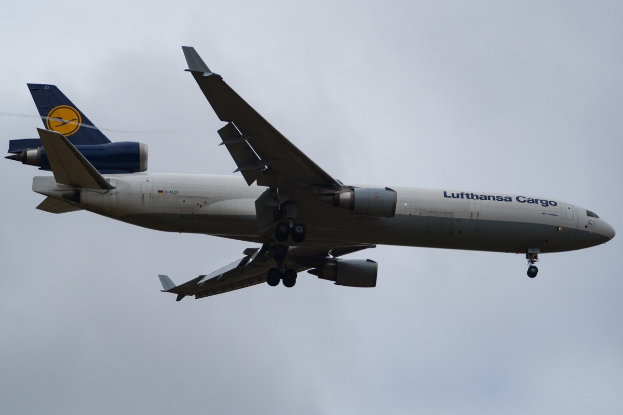 Lufthansa Cargo Boeing 777-300ER im Flug mit sichtbarem Text auf dem Rumpf, ausgefahrenen Flügeln und aktivierten Triebwerken gegen einen Himmel mit weißen Wolken.