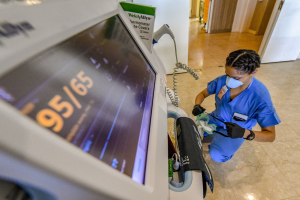 Eine medizinische Fachkraft in Kittel und Mundschutz steht in einem Krankenhausflur und hält ein Gerät, das über einen Draht verbunden ist, mit Türen und medizinischen Geräten im Hintergrund.