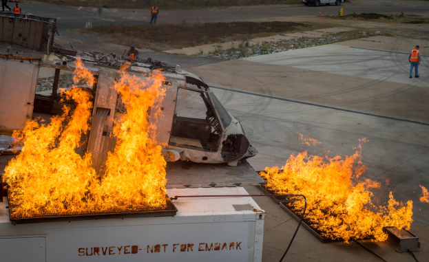 Ein Flugzeug, das auf dem Rollfeld eines Flughafens in Flammen steht, mit einem Fahrzeug links daneben und einigen Menschen in der Nähe, mit weiteren Fahrzeugen und Bäumen im Hintergrund.