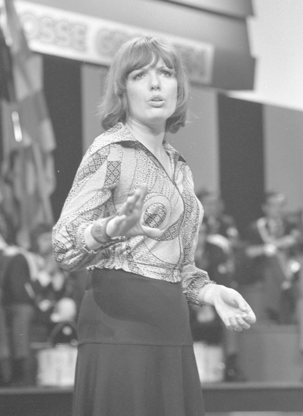 Eine Frau in einem Schwarz-Weiß-Foto steht auf einer Bühne, trägt ein weißes Hemd und einen schwarzen Rock, hat ihre Haare zu einem Dutt gebunden und einen ernsthaften Ausdruck, umgeben von einer Gruppe von Menschen und einer Wand im Hintergrund.