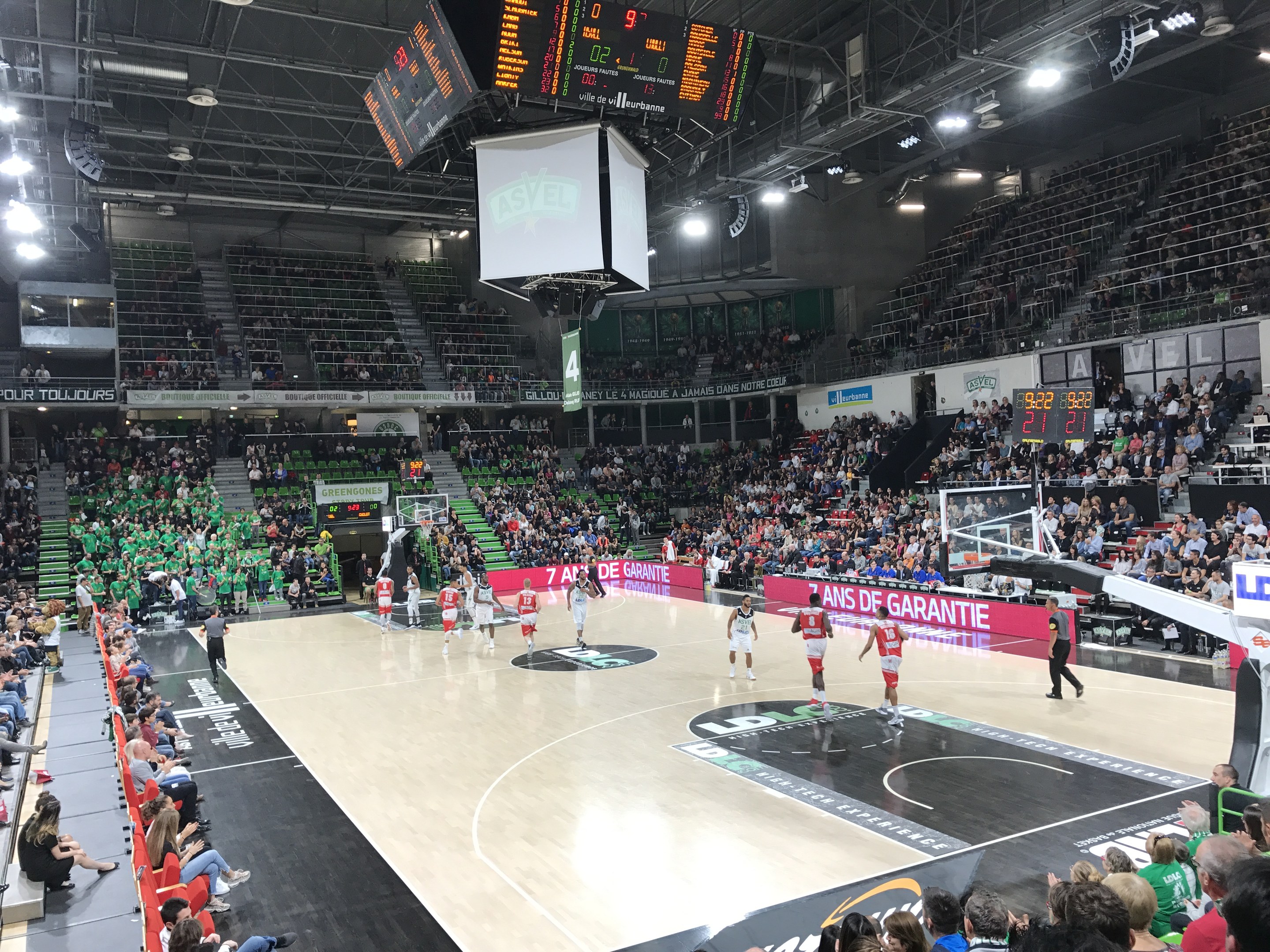 Basketballspiel in einer großen Arena mit Zuschauern, mit Beteiligung der Teams Asvel und Galatasaray, mit Anzeigetafeln und Deckenbeleuchtung.