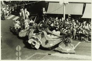 Ein Schwarz-Weiß-Foto eines mit Blumen geschmückten Umzugswagens, mit einer Gruppe von Menschen auf der Straße und einem Pfahl im Hintergrund.