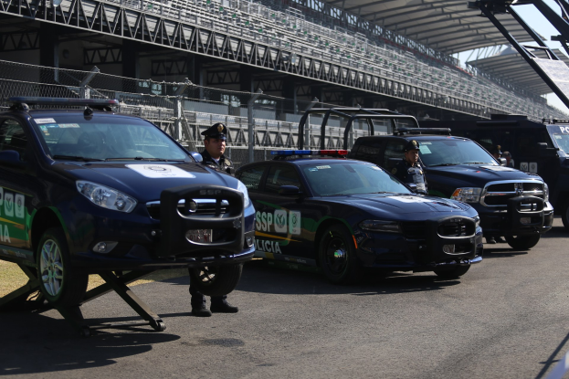 Polizeiautos vor einer Rennstrecke mit uniformierten Personen, ein Stadion im Hintergrund und Maschendraht unter einem sichtbaren Himmel.