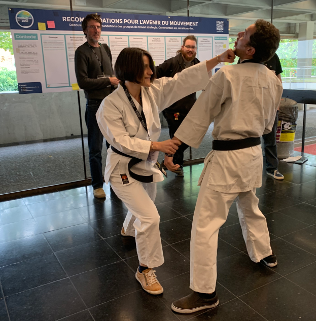 Ein Mann und eine Frau in Karate-Ausrüstung stehen nebeneinander vor einem Plakat mit einer Glaswand dahinter, die Bäume zeigt, und einem Mülleimer und einem Tisch mit Gegenständen rechts daneben, was auf eine Taekwondo-Wettkampfstätte hinweist.