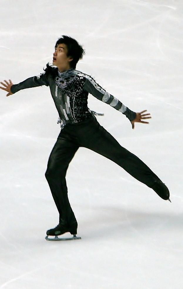 Männlicher Eiskunstläufer gleitet über eine Eisbahn in einem schwarzen Kleid und führt eine Choreografie mit synchronen Arm- und Beinbewegungen aus.