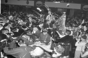 Ein Schwarz-Weiß-Bild einer Menge von Menschen in Schottenröcken, die auf einer Party tanzen, wobei einige Musikinstrumente halten und Texttafeln im Hintergrund zu sehen sind.