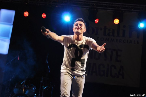 Ein Mann auf einer hell erleuchteten Bühne hält ein Mikrofon in der Hand, im Hintergrund sind Musikinstrumente und ein "Nouveau Magica"-Schild zu sehen.