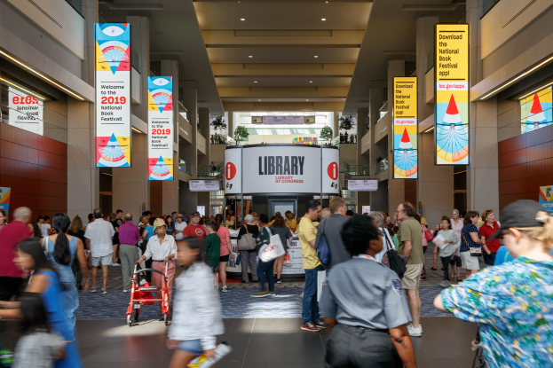 Eine große Gruppe von Menschen, die durch ein Gebäude mit Bannern an Wänden und Säulen geht, einige tragen Taschen und einer schiebt einen Kinderwagen, Deckenlampen und eine Tafel im Hintergrund, die mit dem Nationalen Buchfestival in Verbindung steht.
