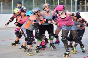 Mehrere Frauen mit Helmen und Schlittschuhschuhen fahren Schlittschuh und schieben sich gegenseitig in der Nähe eines Metallzauns, mit einem Mann und Bäumen im Hintergrund.