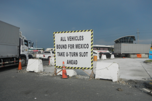 Ein Lastwagen auf einem Parkplatz neben einem Schild mit der Aufschrift "Alle Fahrzeuge nach Mexiko", mit verstreuten Gegenständen, Gebäuden, Bäumen, Pfählen und einem klaren blauen Himmel im Hintergrund.