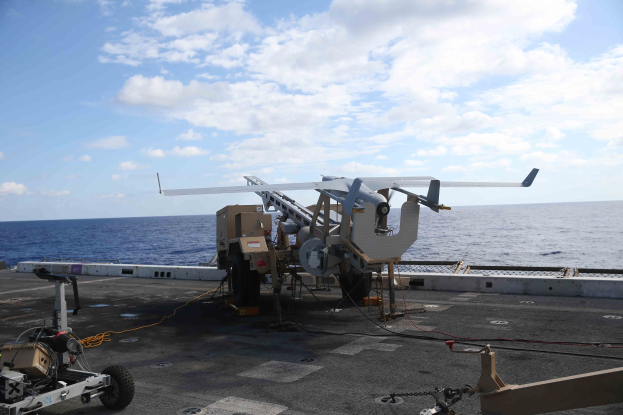 Unbemanntes Luftfahrzeug (UAV) auf dem Deck eines Flugzeugträgers mit Meer und wolkenverhangenem Himmel im Hintergrund.