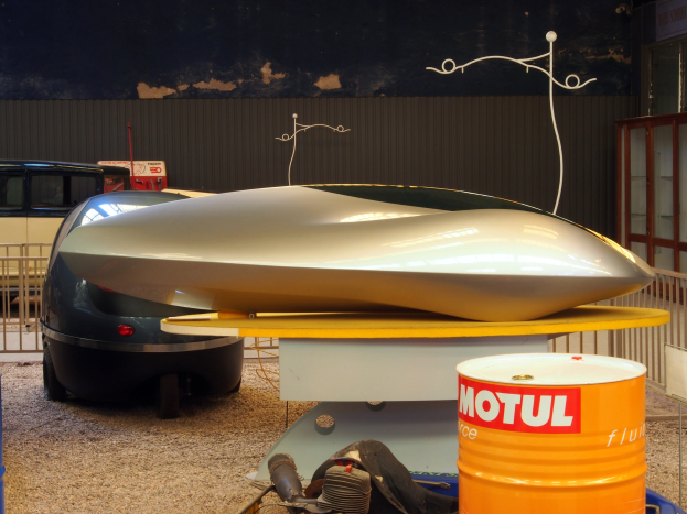 Kleines Hydrofoil-Boot auf einem Tisch neben einem Auto, mit zwei Trommeln auf dem Boden, einem Metallzaun im Hintergrund und einer Glastür mit einer Texttafel auf der rechten Seite.