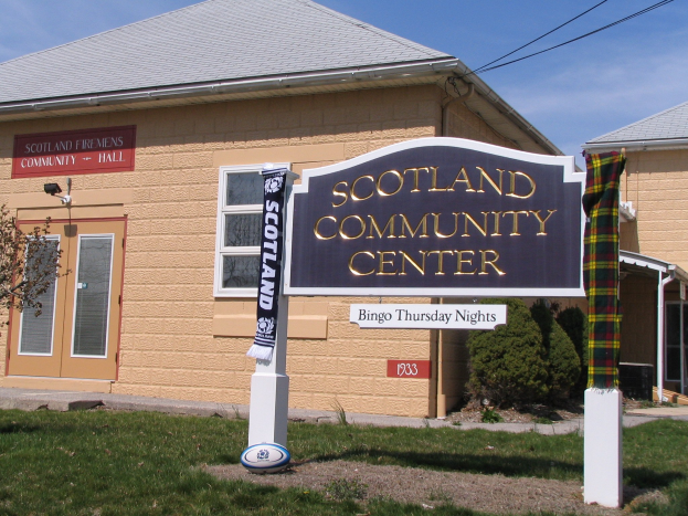 Außenansicht des Scotland Community Center mit einem Namen-Schild, Gras, einem Ball, Pflanzen, einem Baum, Überlandleitungen und einem bewölkten Himmel.