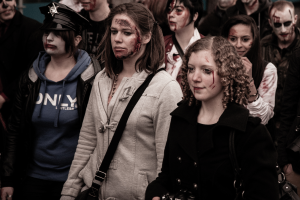 Eine Gruppe von Menschen in Zombie-Kostümen, einige in Verkleidungen, einer mit Hut und Handtasche, die eine Straße entlanggehen mit einem Gebäude im Hintergrund.