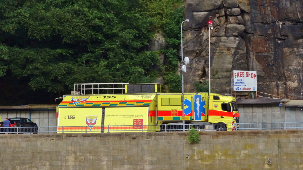 Ambulanz neben einer Klippe geparkt, mit verschiedenen Objekten im Vordergrund und Bäumen und einem Auto im Hintergrund.