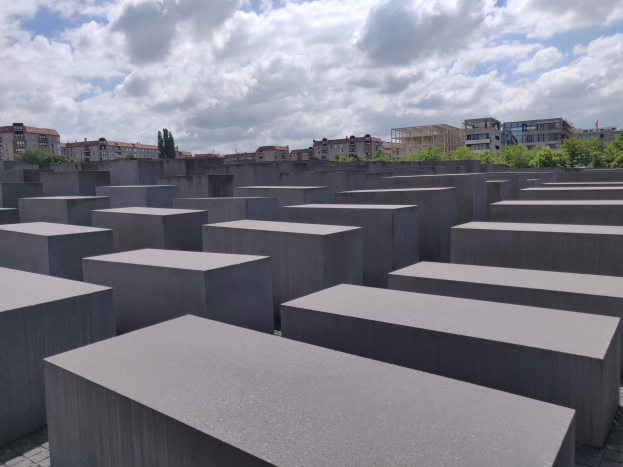 Betonschirme in einem Gittermuster auf dem Holocaust-Mahnmal für die ermordeten Juden Europas in Berlin mit Bäumen, Gebäuden und Wolken im Hintergrund.