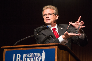 Ein Mann in Anzug und Krawatte steht an einem Podium und hält eine Auszeichnung, mit einem Mikrofon und einer Tafel am Podium, während einer Bibliothekspreisverleihung.